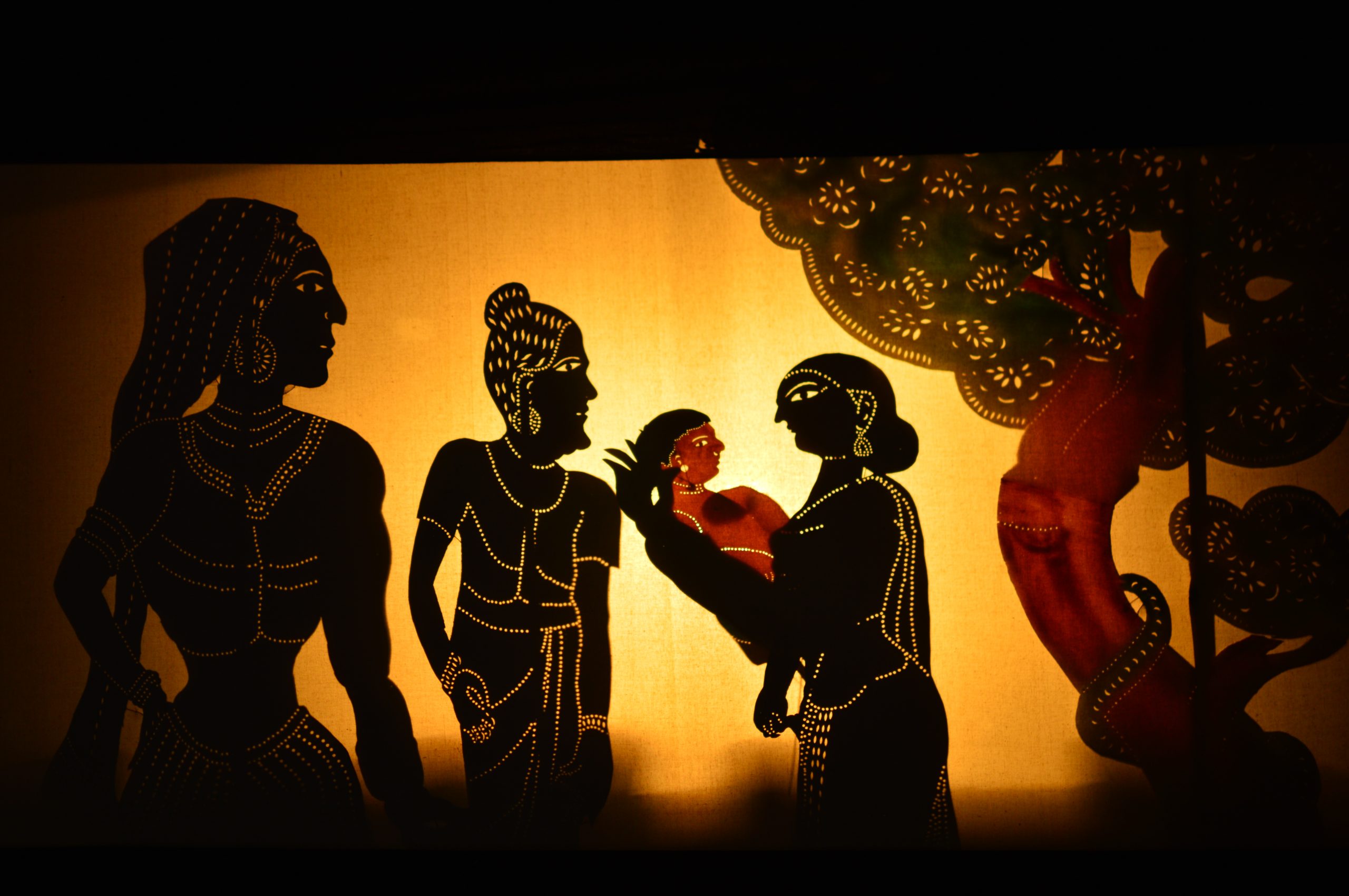 Image portrays shadow puppetry in action. Four figures, one a child, are grouped together beside a stylized tree.