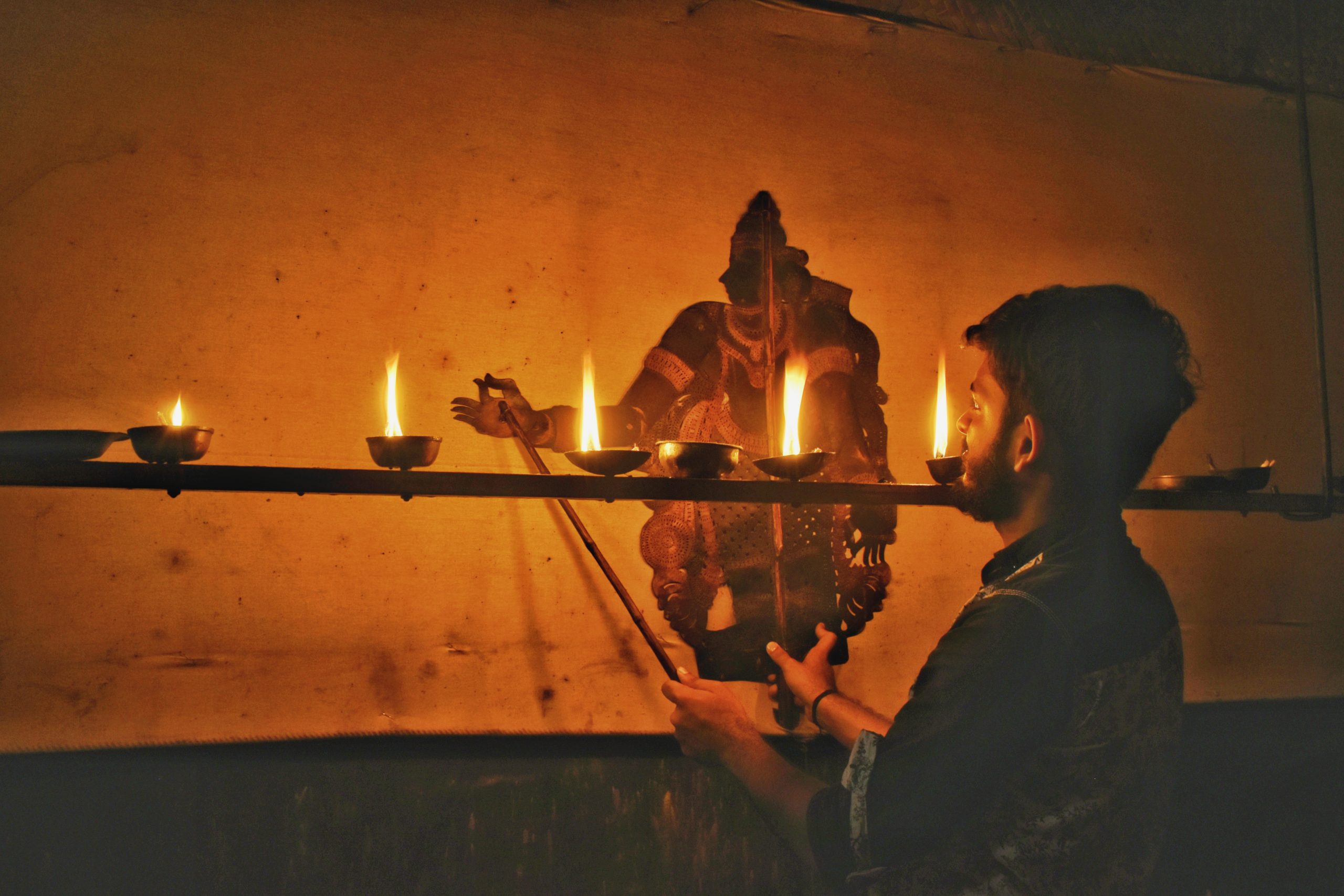 Image portrays traditional puppet manipulation onto screen lit by candles. Rahul Koonathara is manipulating the shadow puppet.